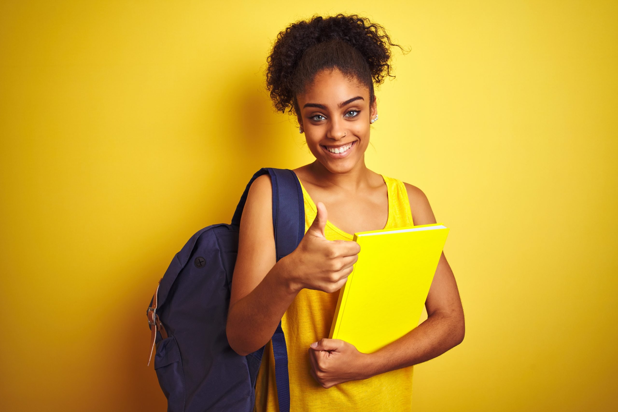 A cheerful young woman with curly hair giving a thumbs-up while holding a bright yellow notebook, wearing a yellow tank top and a blue backpack, standing against a vivid yellow background, attesting to the success gained with Canberra Tutoring.